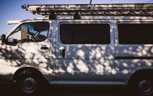 White utility van with a roof ladder rack parked, shadows from trees on its side.