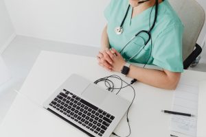 Healthcare professional in scrubs with stethoscope sits at a white desk, hands folded, facing a laptop on a clean workspace.