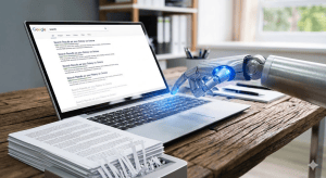 Robotic hand typing on a laptop displaying search results, with a stack of papers on a wooden desk.